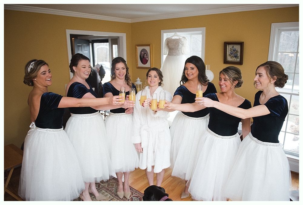 Bridesmaids in red dresses and shawls laugh with bride holding a bouquet outdoors.