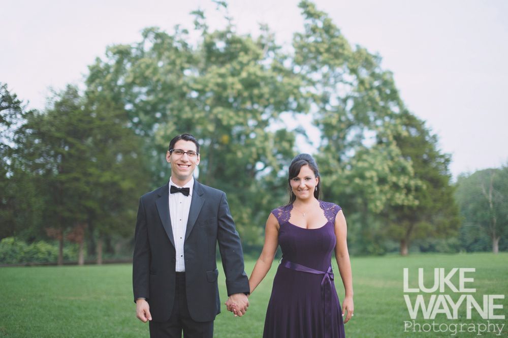 Couple holding hands, smiling, in formal attire, standing on grass with trees in the background.