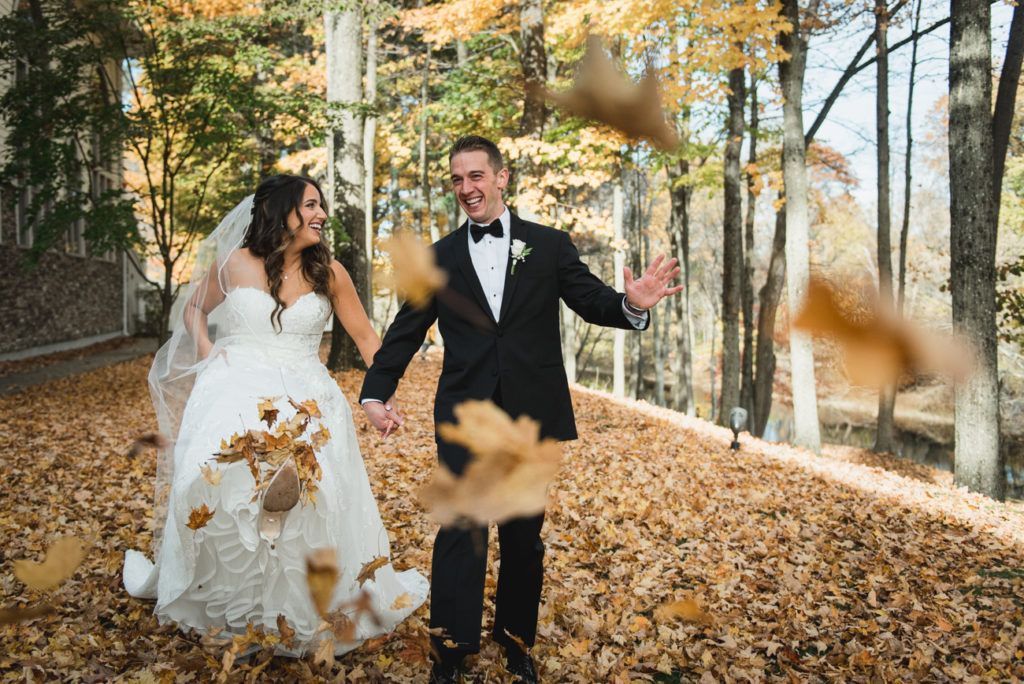 Bride and groom celebrating among falling autumn leaves; trees and foliage in background.