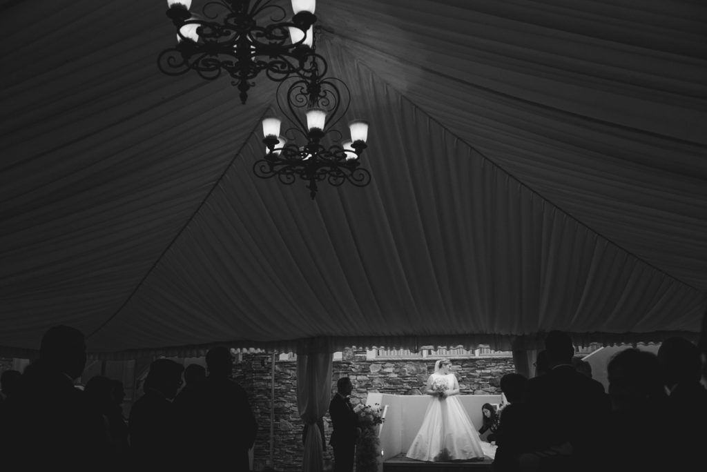 Bride in a wedding dress under a tent with chandeliers; guests watch.
