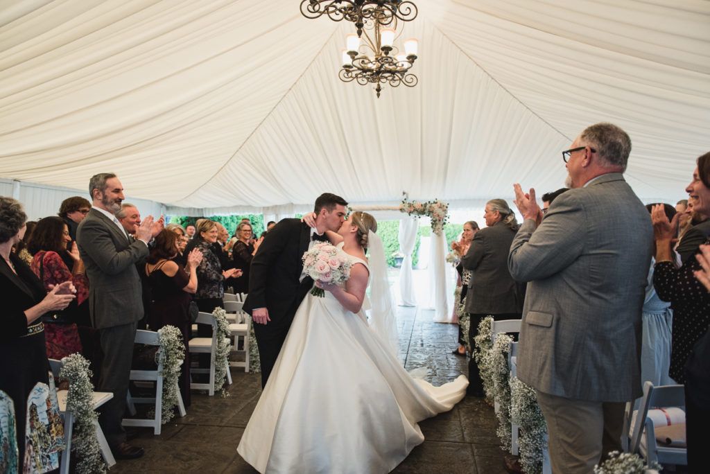 Bride and groom kiss at wedding ceremony, guests clap, tent setting, white gown, black suit.