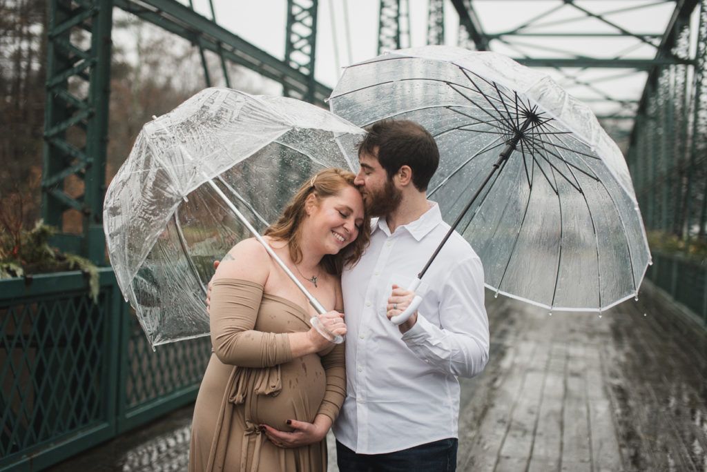 Couple under clear umbrellas on a bridge, woman pregnant, man kisses her forehead, outdoors.