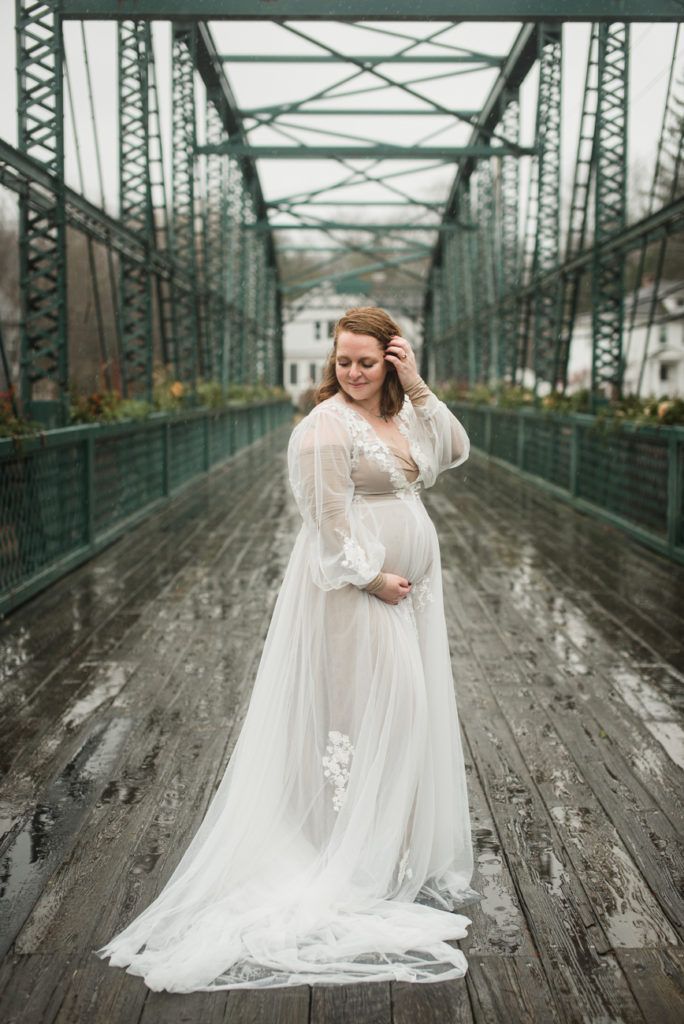 Pregnant woman in white gown on a bridge, smiling, hand on belly.