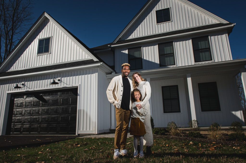 Family of three standing in front of a white house with a black garage door.