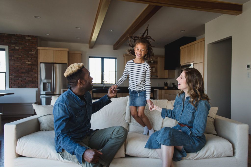 A family smiles, a child jumping between parents on a cream sofa in a living room.