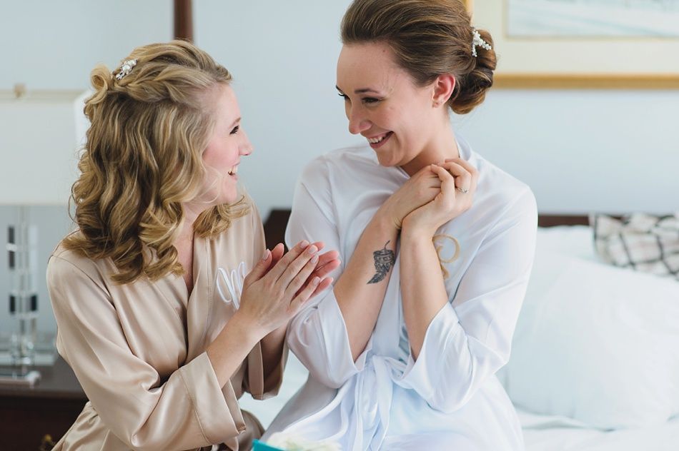 Wedding party on a beach with blue sky. Bride and groom embrace, others stand nearby. Dark blue dresses and jackets.