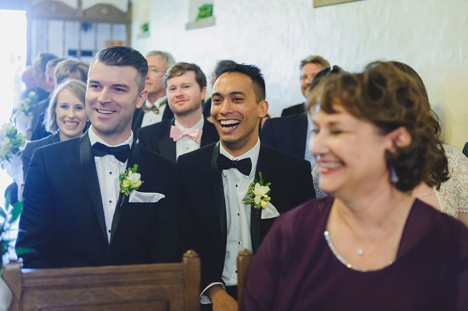 Bride with arms up, making a funny face, groom looking at his hand, cake cutting in background.