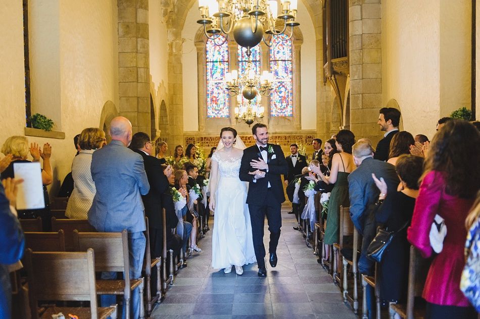 Bride with arms up, making a funny face, groom looking at his hand, cake cutting in background.