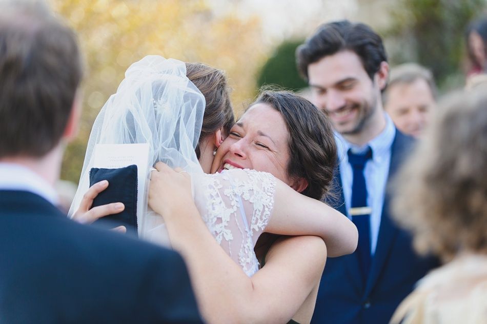 Bride with arms up, making a funny face, groom looking at his hand, cake cutting in background.