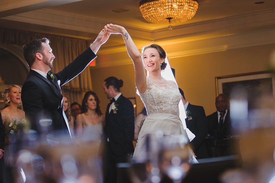 Bride with arms up, making a funny face, groom looking at his hand, cake cutting in background.