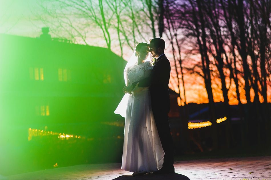 Bride with arms up, making a funny face, groom looking at his hand, cake cutting in background.