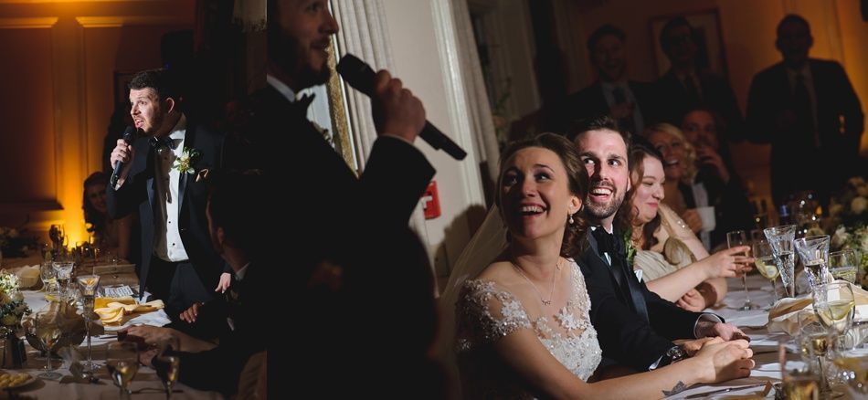 Bride with arms up, making a funny face, groom looking at his hand, cake cutting in background.
