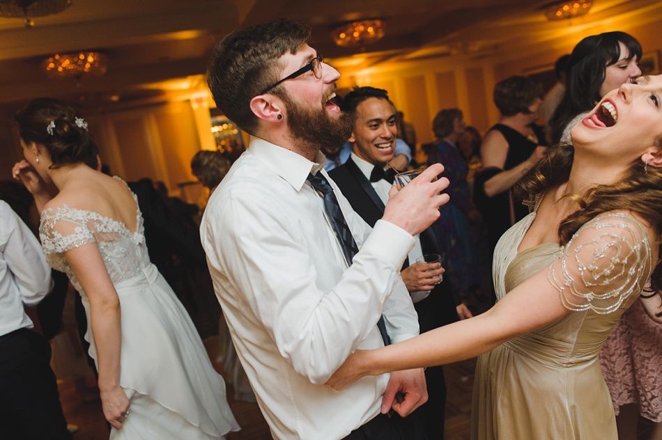 Bride with arms up, making a funny face, groom looking at his hand, cake cutting in background.