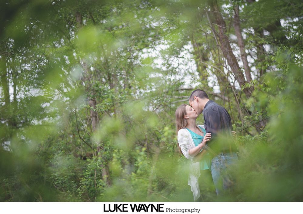 Couple kissing in a forest, framed by green foliage; the man wears a dark shirt, woman has a teal top and a white shawl.