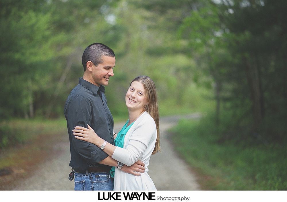 Couple embracing on a path in a wooded area, smiling at each other.