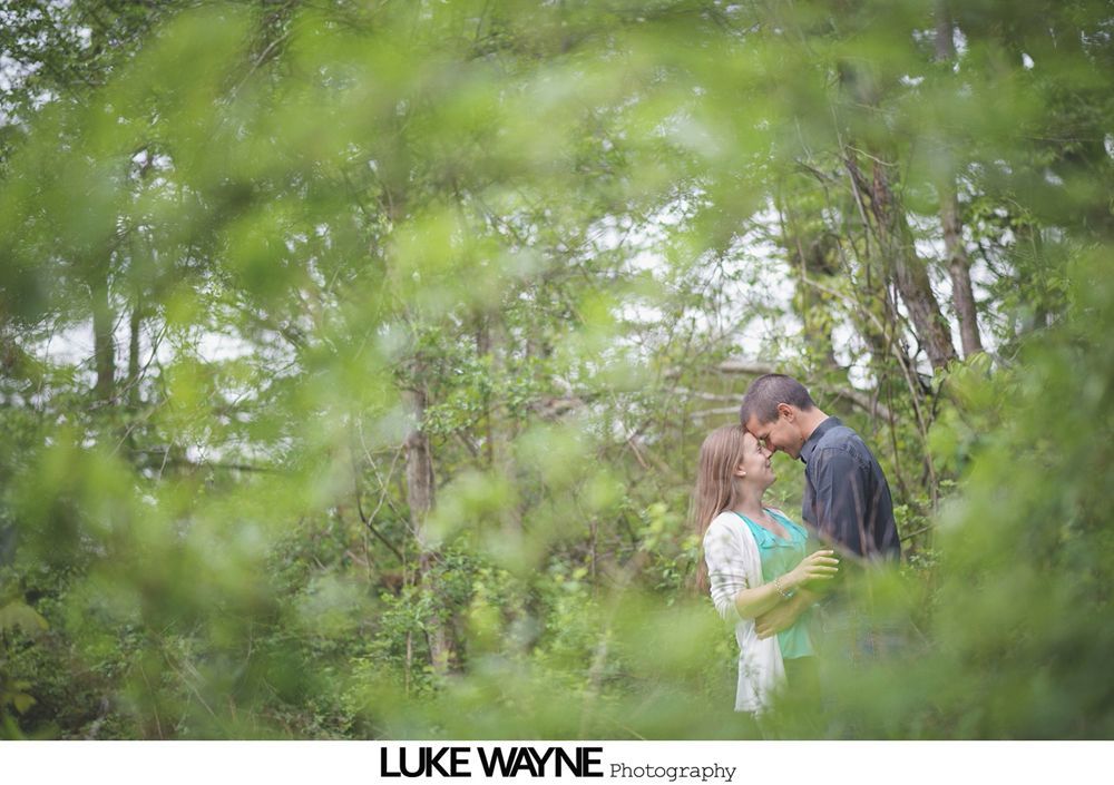 Couple embracing in a forest, surrounded by green foliage.