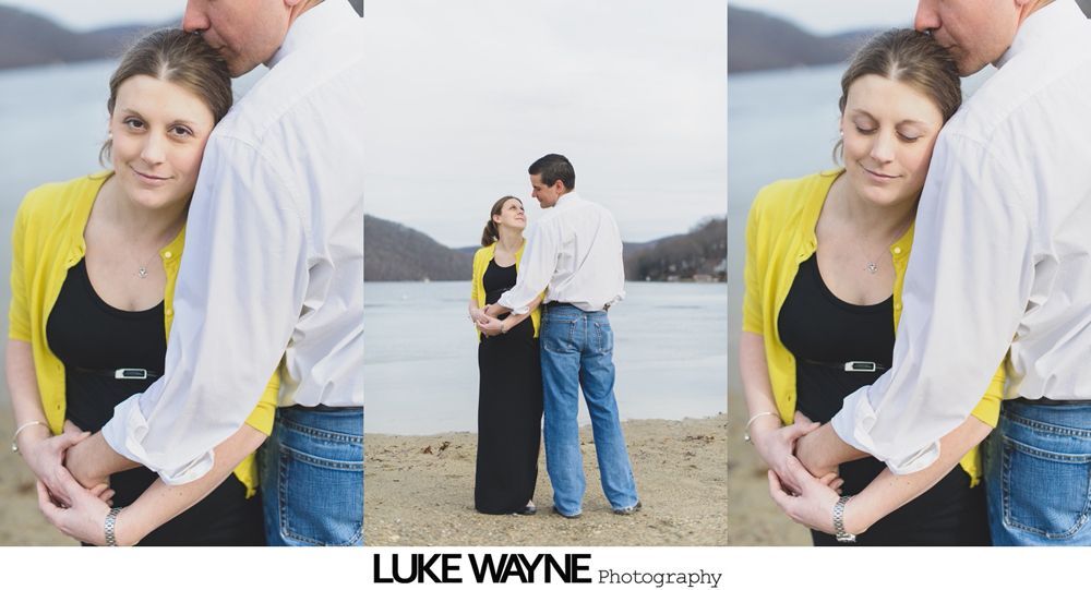 Couple embraces on a beach, woman in yellow cardigan, man kisses her forehead, lake in background.