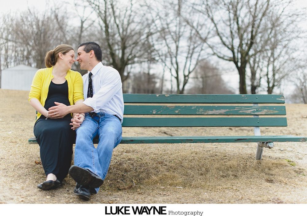 Couple sits on park bench, holding hands, smiling. Woman's pregnant belly visible.