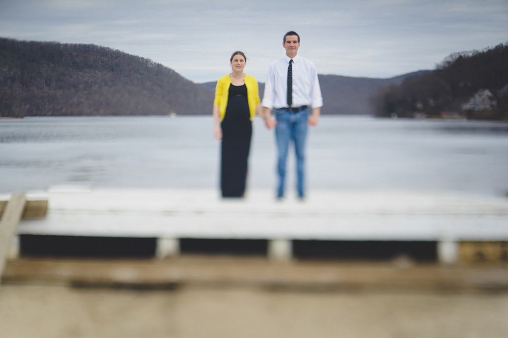Couple stands holding hands on a dock; woman in yellow sweater and black dress, man in white shirt and jeans; lake and hills background.