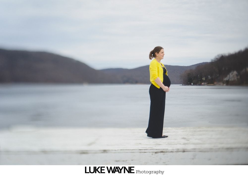 Pregnant woman stands by a frozen lake, wearing a yellow sweater and black dress, mountains in background.
