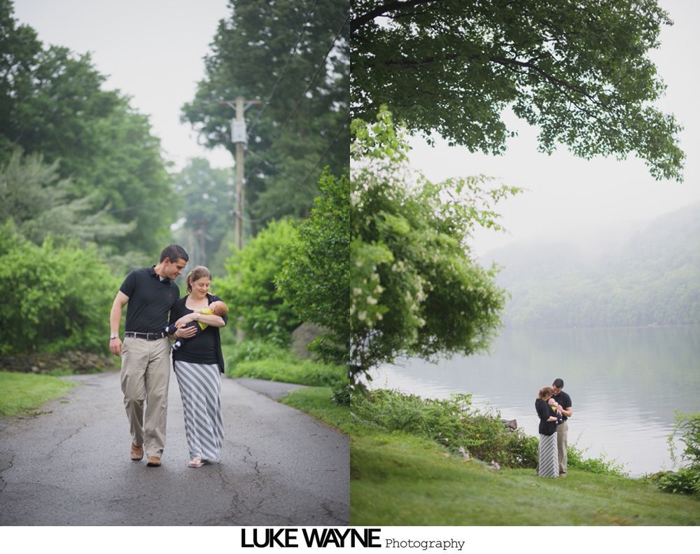 Couple walking and hugging by a lake, holding a baby. Overcast, lush greenery.
