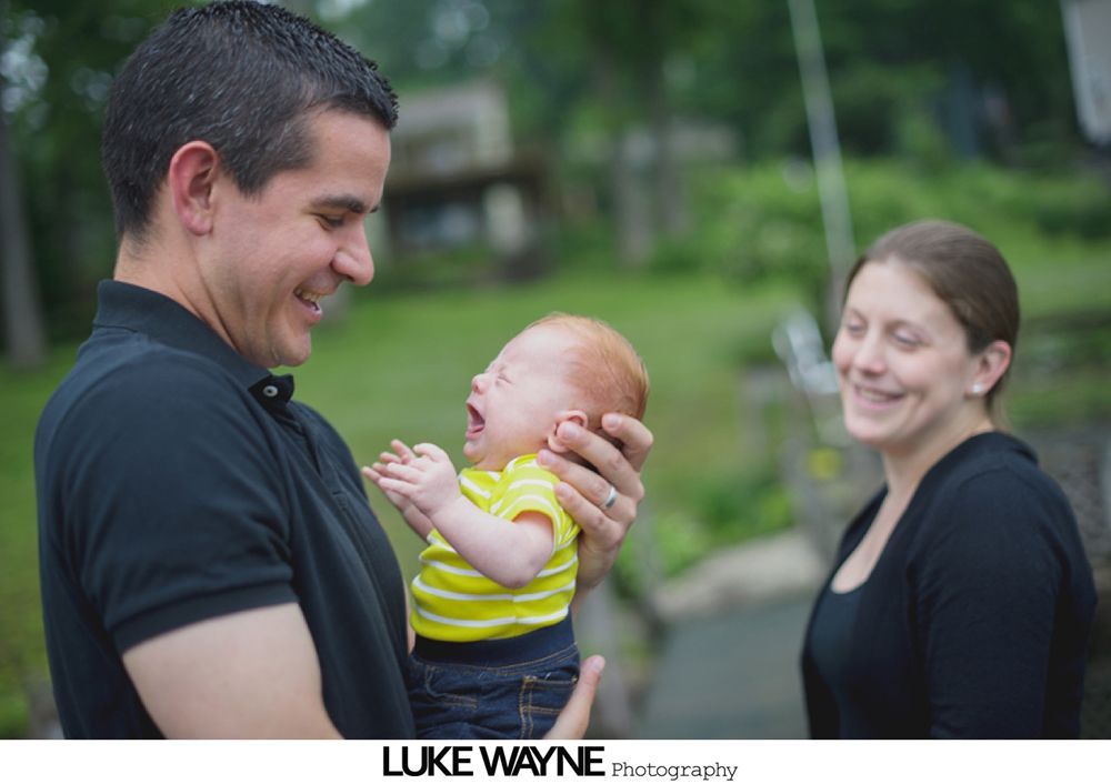 Man holding laughing baby, woman smiles in background; outdoor setting.