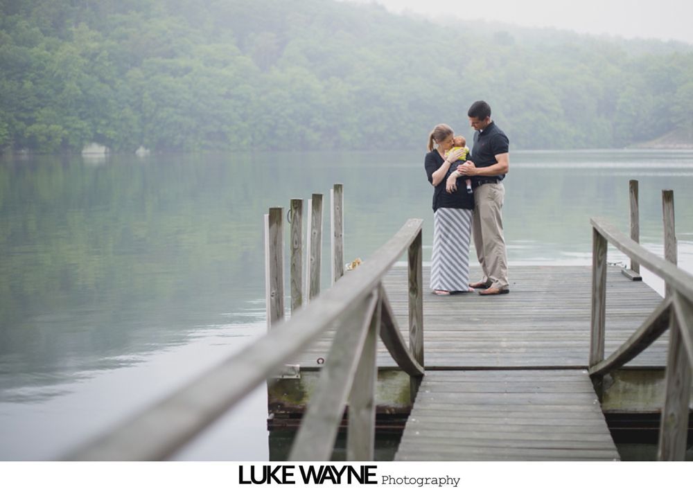 Couple on a dock holding a baby, overlooking a calm lake on a misty day.