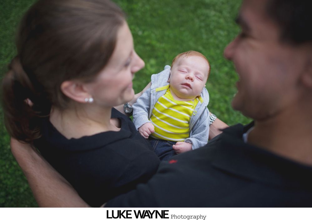 Parents holding sleeping baby, smiling. Green grass background.