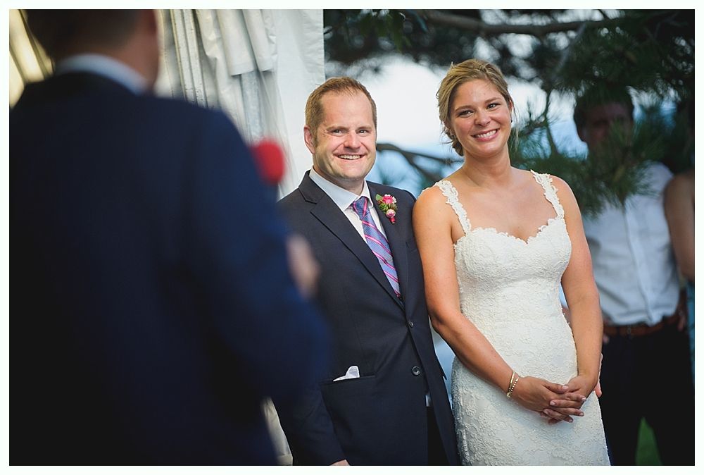 Bride with arms up, making a funny face, groom looking at his hand, cake cutting in background.