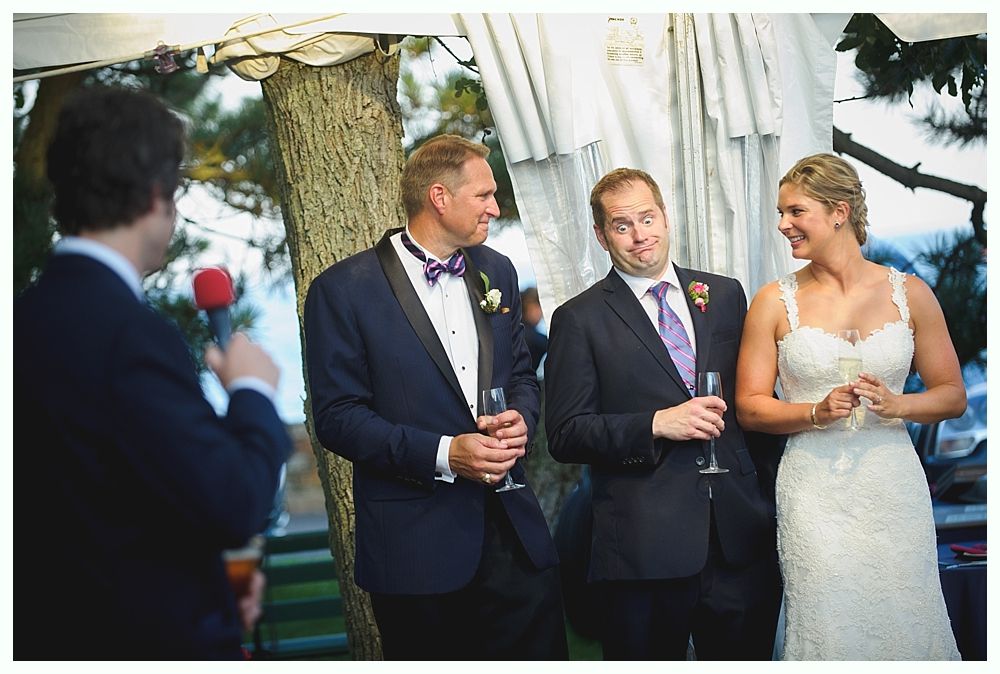 Bride with arms up, making a funny face, groom looking at his hand, cake cutting in background.