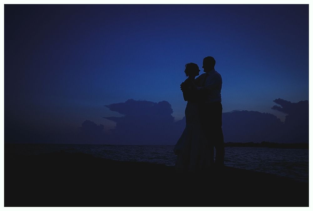 Bride with arms up, making a funny face, groom looking at his hand, cake cutting in background.