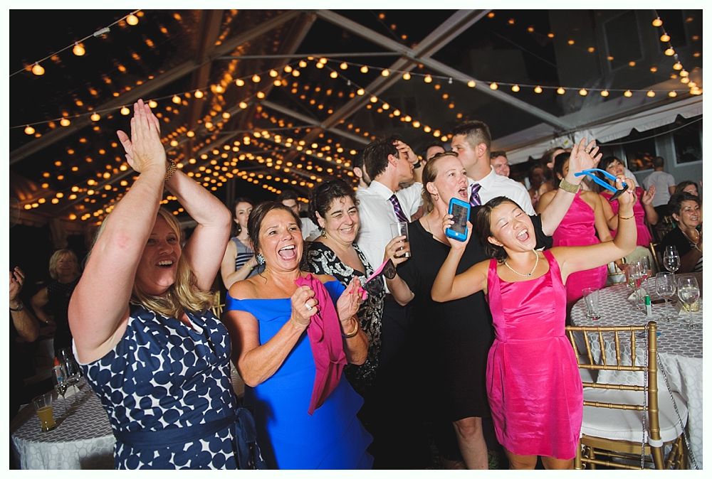 Bride with arms up, making a funny face, groom looking at his hand, cake cutting in background.