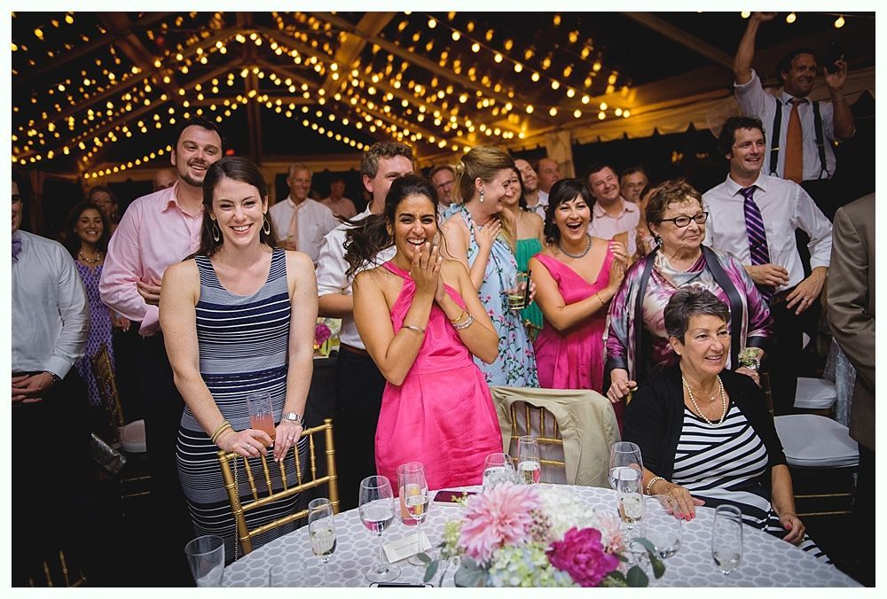 Bride with arms up, making a funny face, groom looking at his hand, cake cutting in background.