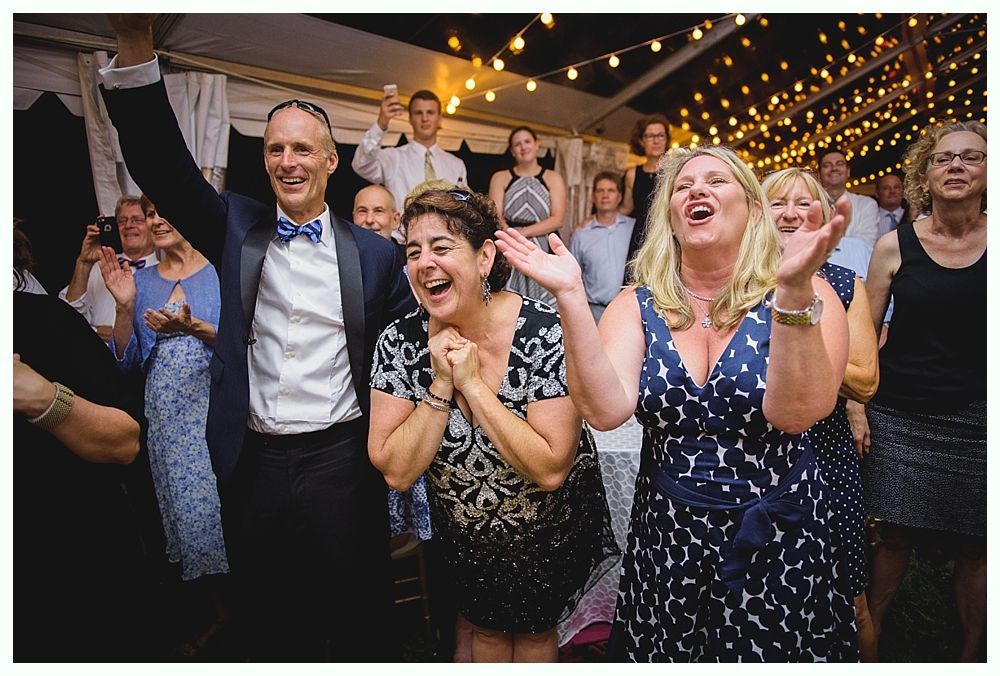 Bride with arms up, making a funny face, groom looking at his hand, cake cutting in background.
