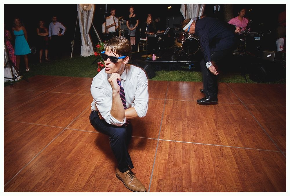 Bride with arms up, making a funny face, groom looking at his hand, cake cutting in background.