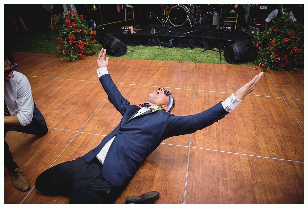 Bride with arms up, making a funny face, groom looking at his hand, cake cutting in background.