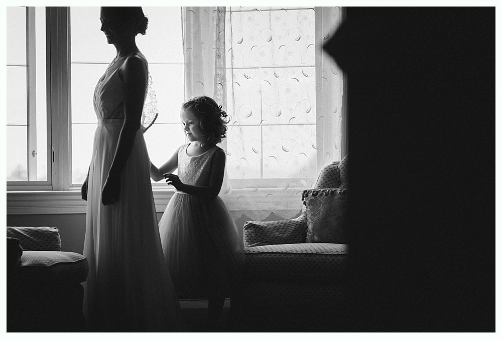 Bride and groom embrace on rocks by the ocean at sunset. The groom wears a blue suit, the bride a white dress.