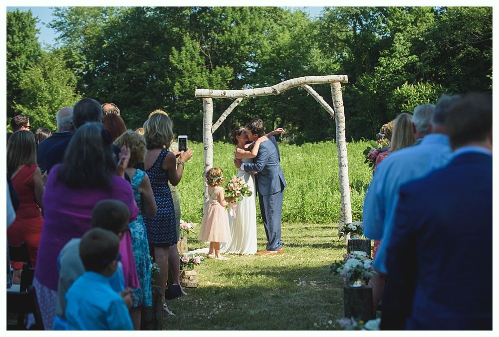 Bride with arms up, making a funny face, groom looking at his hand, cake cutting in background.
