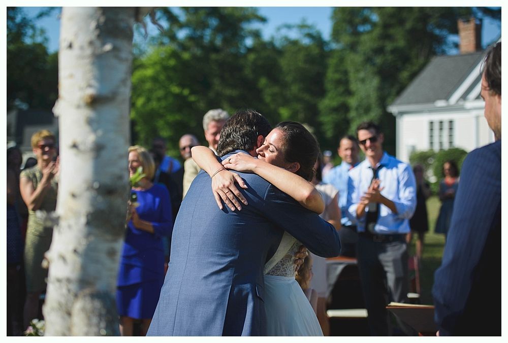 Bride with arms up, making a funny face, groom looking at his hand, cake cutting in background.