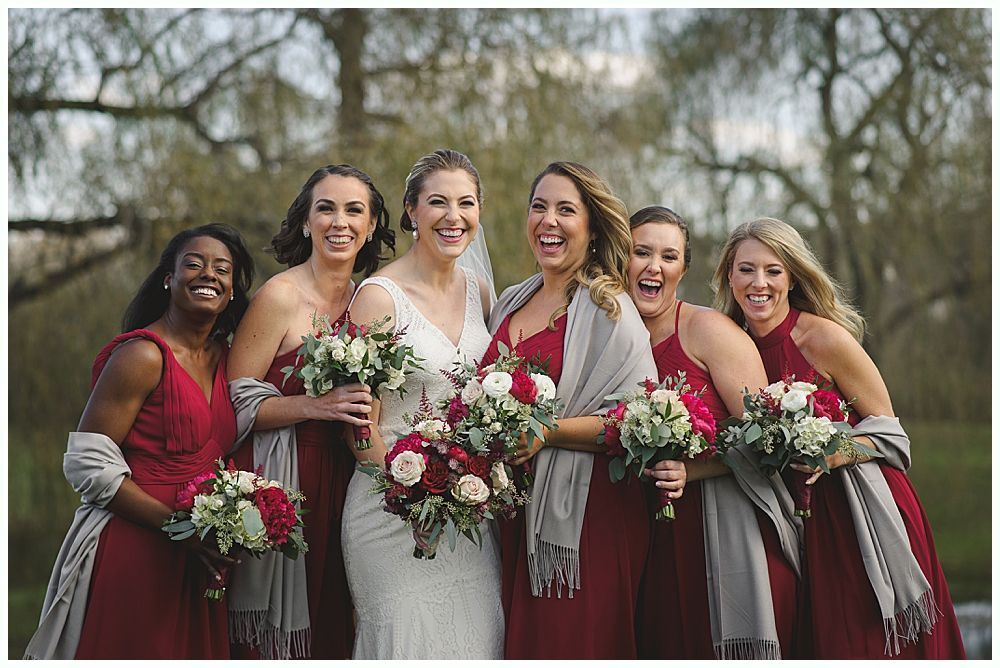 Bridesmaids in red dresses and shawls laugh with bride holding a bouquet outdoors.