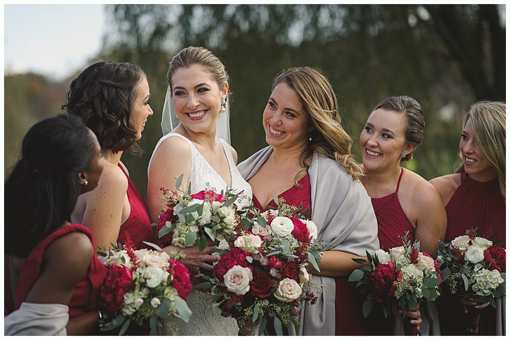 Bride in white dress with bridesmaids in red holding bouquets, smiling outdoors.