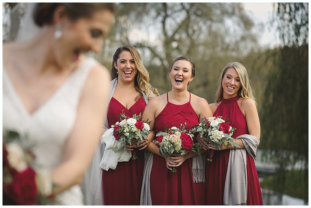 Bride laughs with three bridesmaids in red dresses, holding bouquets.