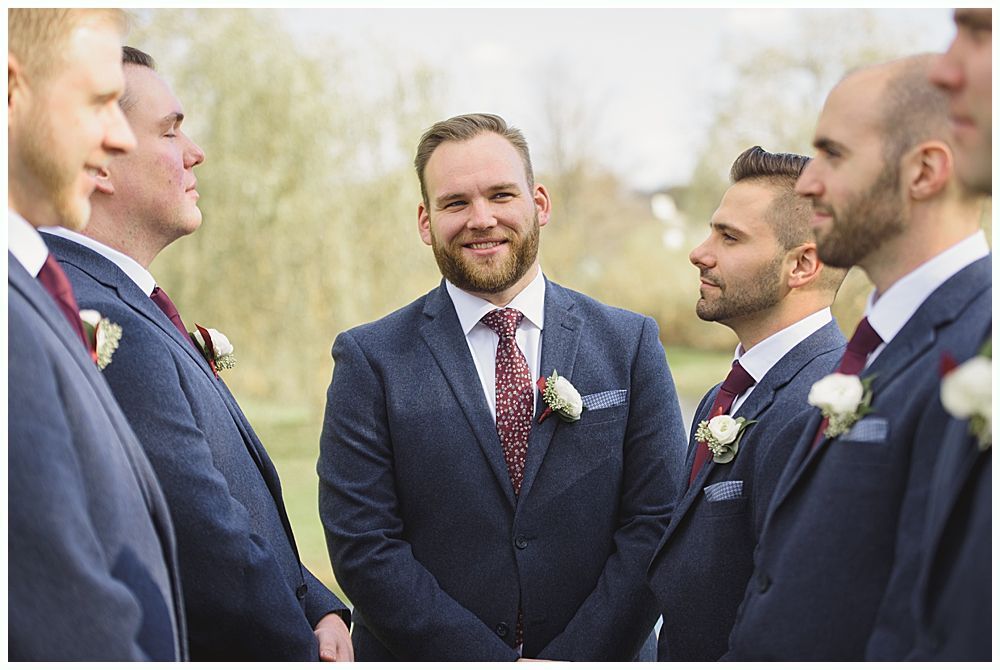 Groom smiles at camera, surrounded by groomsmen in navy suits and burgundy ties, outdoors.
