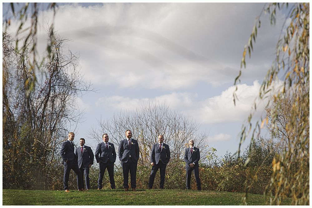 Groomsmen in dark suits pose on a grassy hillside under a cloudy sky.