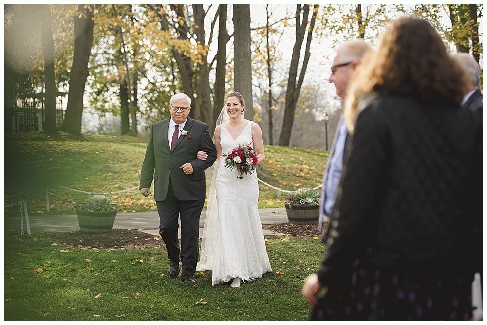 Bride walking with her father down an outdoor aisle; fall trees and guests in background.