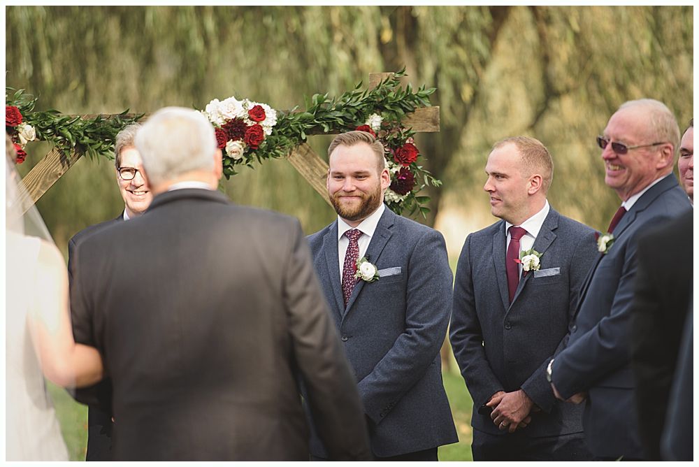 Wedding ceremony with groom and groomsmen smiling, floral arch in background.