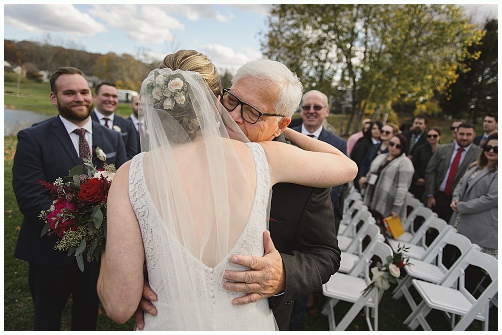 Bride hugging a man near outdoor wedding seating. People watch. The bride wears a white dress and veil.