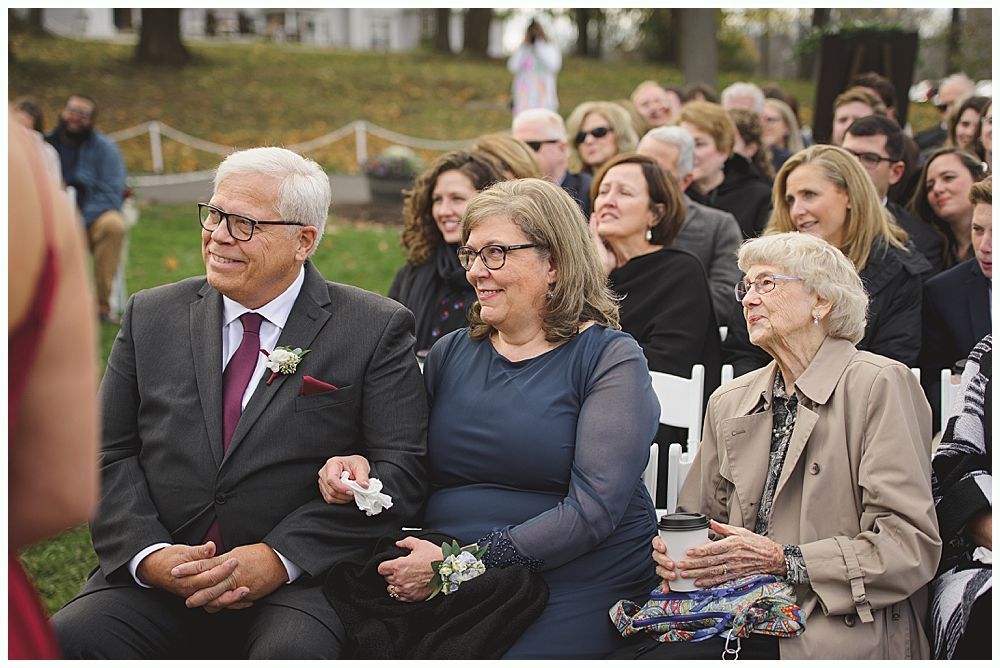 People seated at outdoor ceremony. Elderly woman in beige coat, couple in formal attire, others in background.