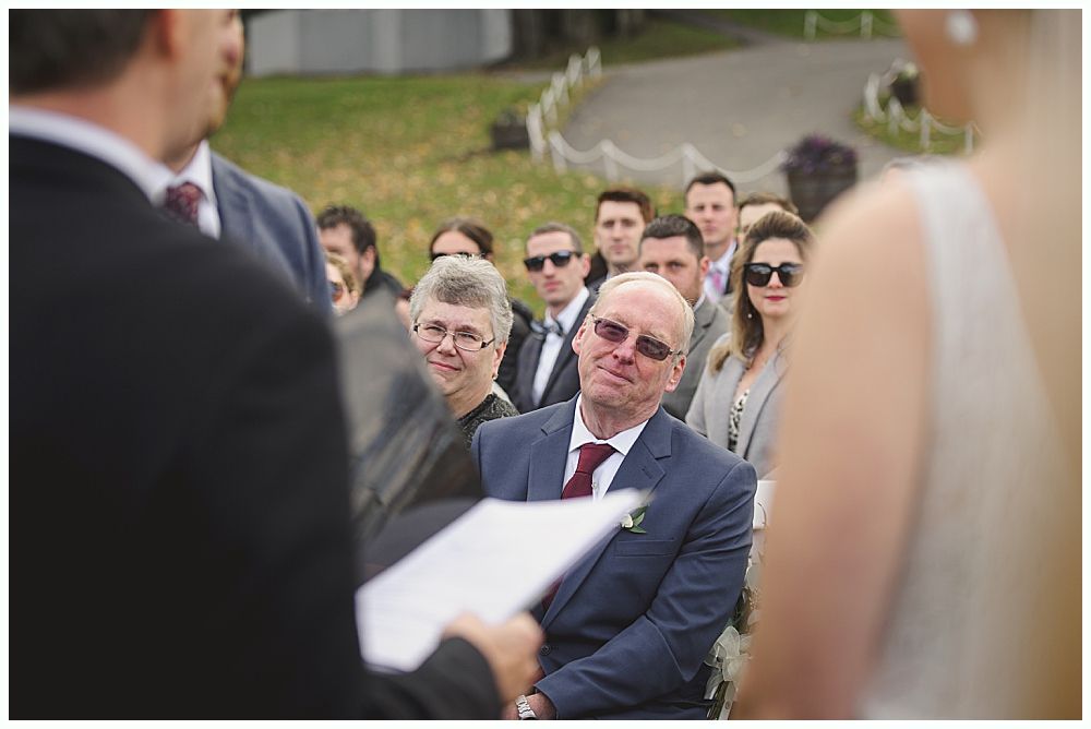 Wedding ceremony. Guests seated, smiling. Groom facing officiant. Bride in white dress. Outdoors, sunny day.