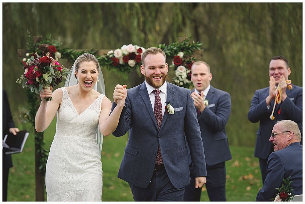 Newlyweds exiting outdoor wedding ceremony, holding hands, smiling. Wedding party applauds.